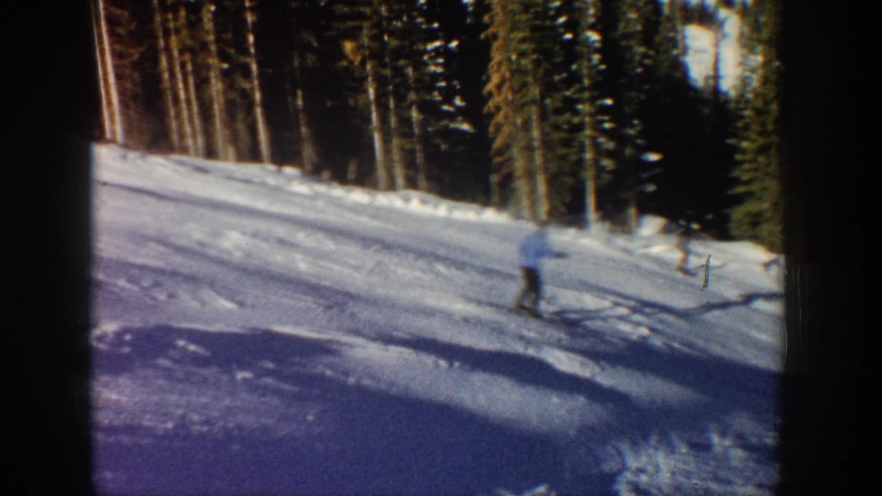 A skier descends a sunlit snowy run in Aspen, Colorado, with tall evergreens and dark film borders visible in the 1961 home-movie frame.