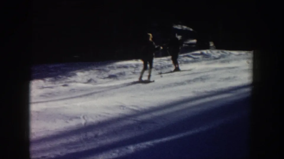 Two dark skier figures appear on a shadowed Aspen snow slope, with strong contrast and film-edge darkness from the 8mm scan.