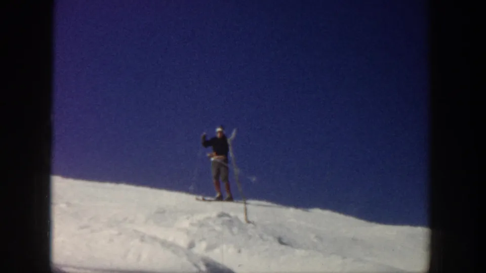 A skier in dark upper clothing stands with poles on an Aspen snow ridge, framed by clear blue sky and 8mm film texture.
