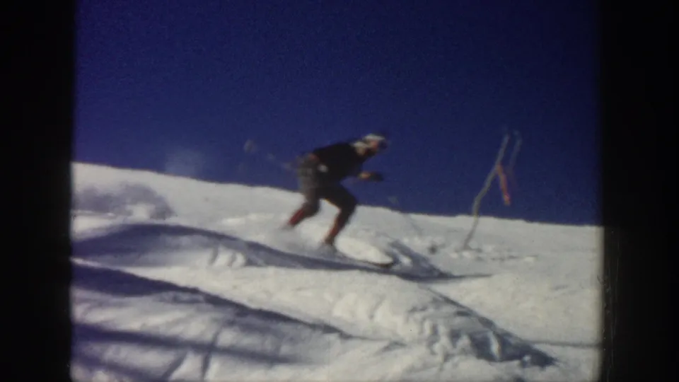 A skier leans forward while descending an uneven Aspen slope, with poles and thin route markers visible in the snow.