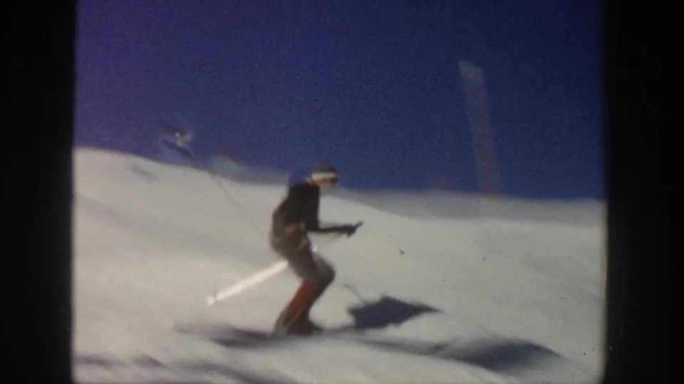The skier crouches low during a downhill pass on a snowy Aspen slope, with a strong shadow and blue sky behind.