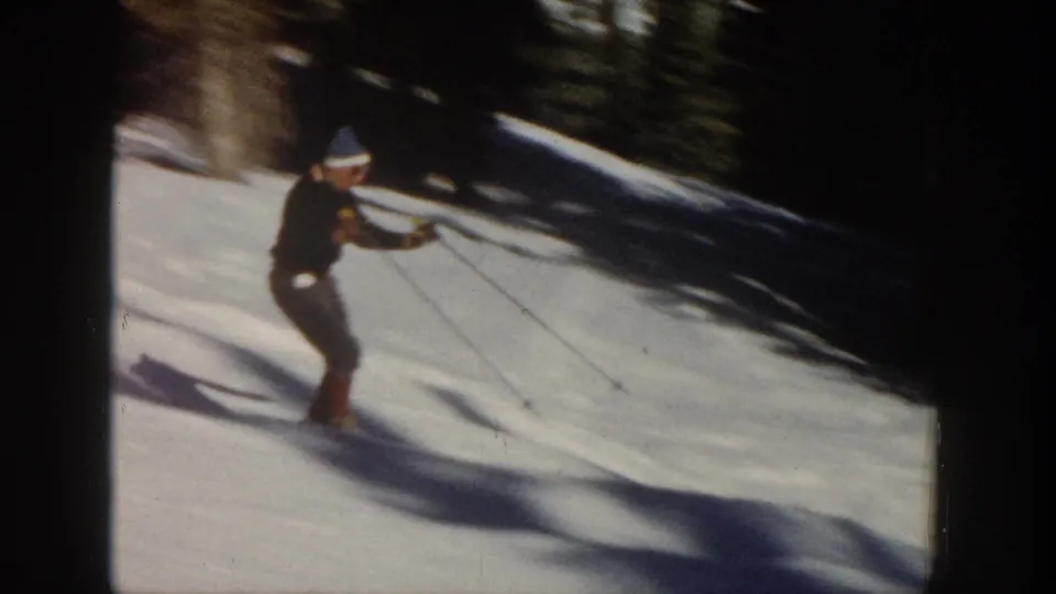 1961 Aspen home-movie frame showing the skier leaning into motion on a snowy run with poles extended and trees behind.