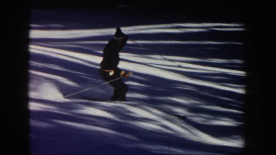 1961 Aspen home-movie frame showing a skier crossing shadowed snow, with strong contrast from trees or terrain outside the frame.