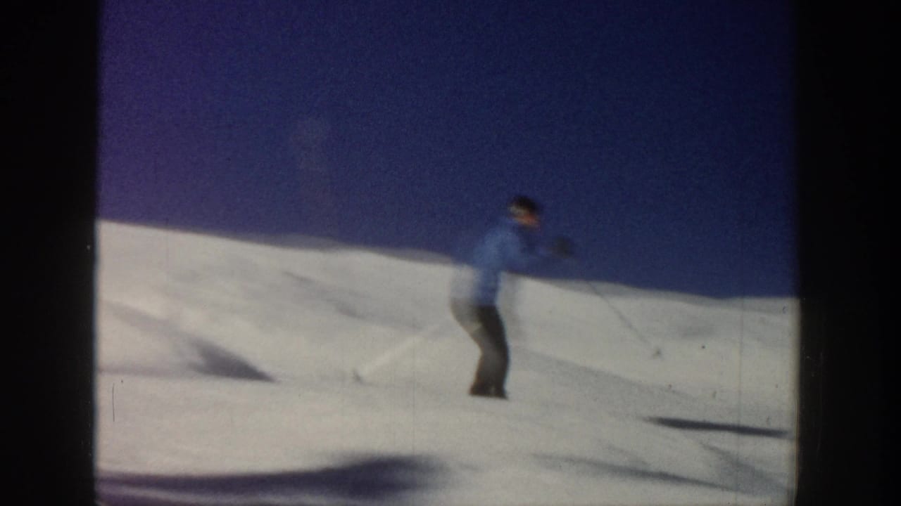 1961 Aspen home-movie frame of a skier in a blue jacket traversing a sunlit snowy slope beneath a clear blue sky.