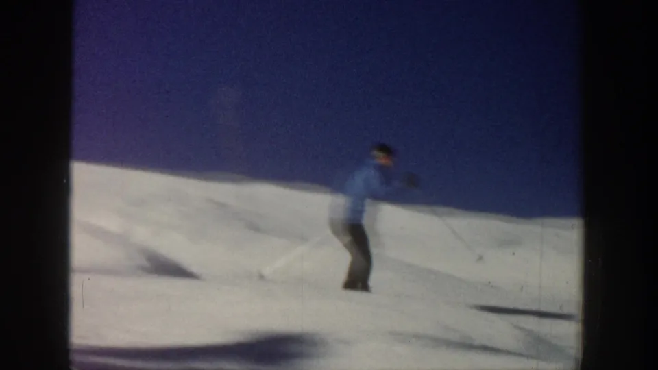 1961 Aspen home-movie frame of a skier in a blue jacket traversing a sunlit snowy slope beneath a clear blue sky.