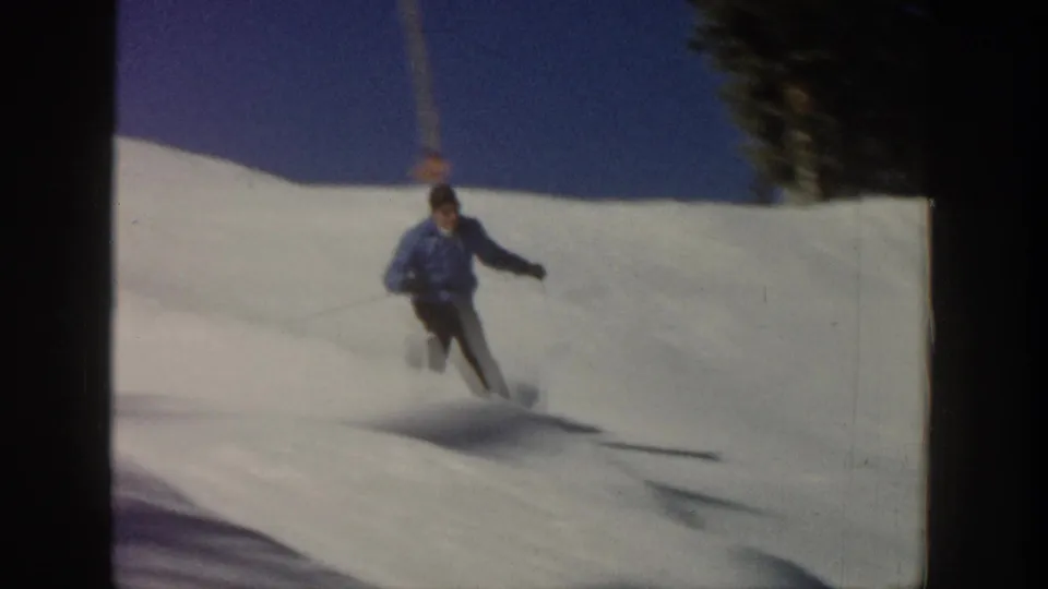 1961 Aspen home-movie frame showing the skier descending through soft focus on a bright slope, with trees near the upper edge.