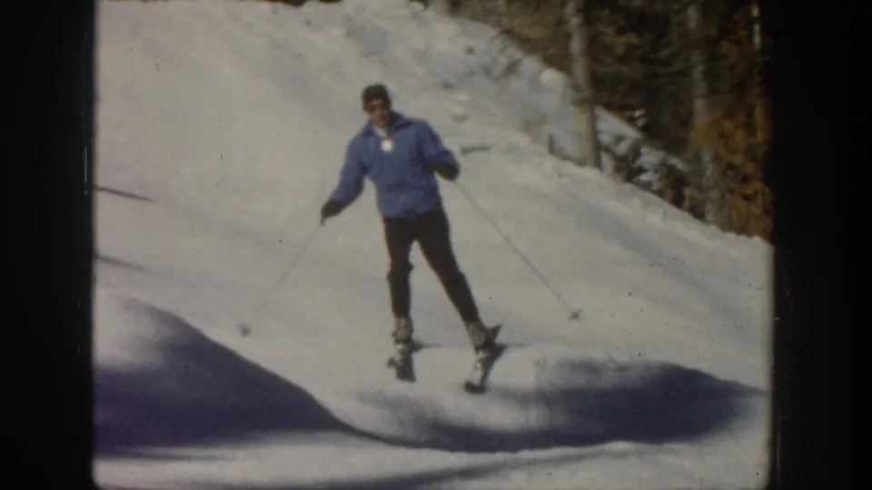 1961 Aspen home-movie frame of a skier in a blue jacket descending near a tree-lined edge of the snowy slope.