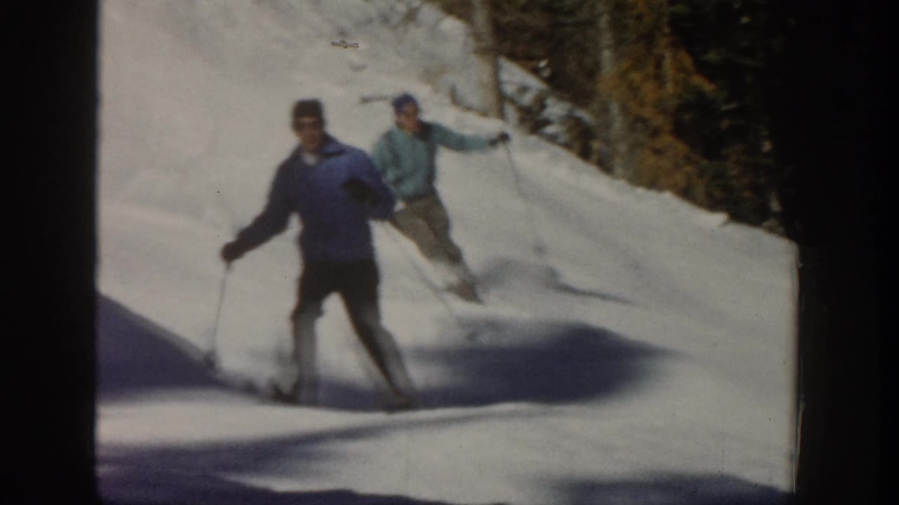 Two adult men ski down a sunny Aspen, Colorado slope in 1961 home-movie footage, with trees along the snowbank.