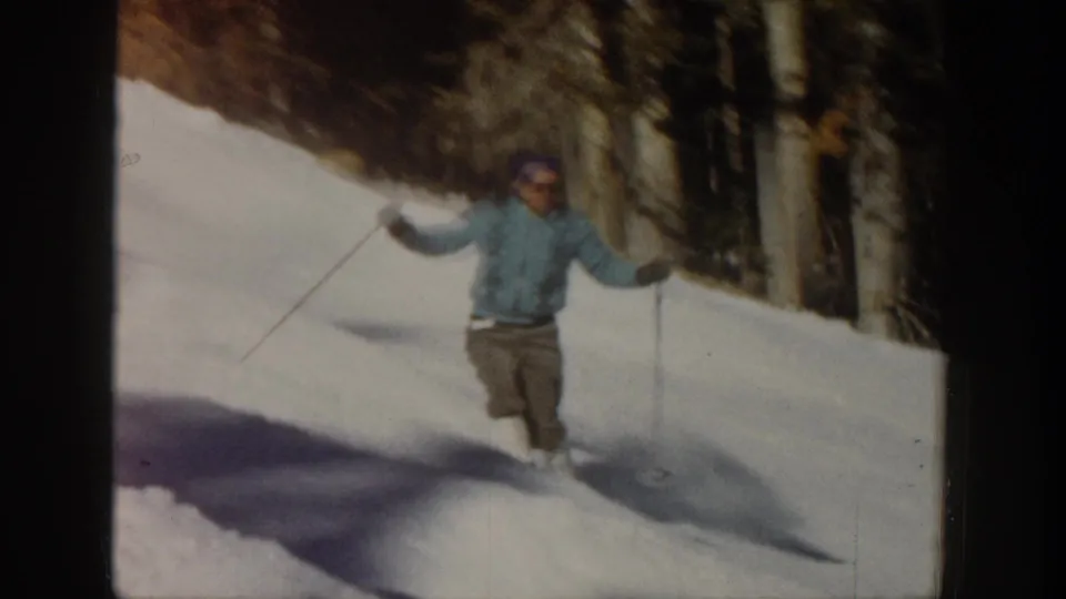 A skier in a light blue jacket descends a snowy Aspen slope with ski poles, framed by dark trees in 1961 8mm footage.