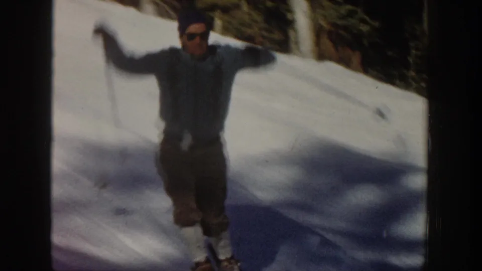 An adult skier approaches the camera on a sunny Aspen snow slope, arms spread with ski poles in a close 8mm home-movie frame.