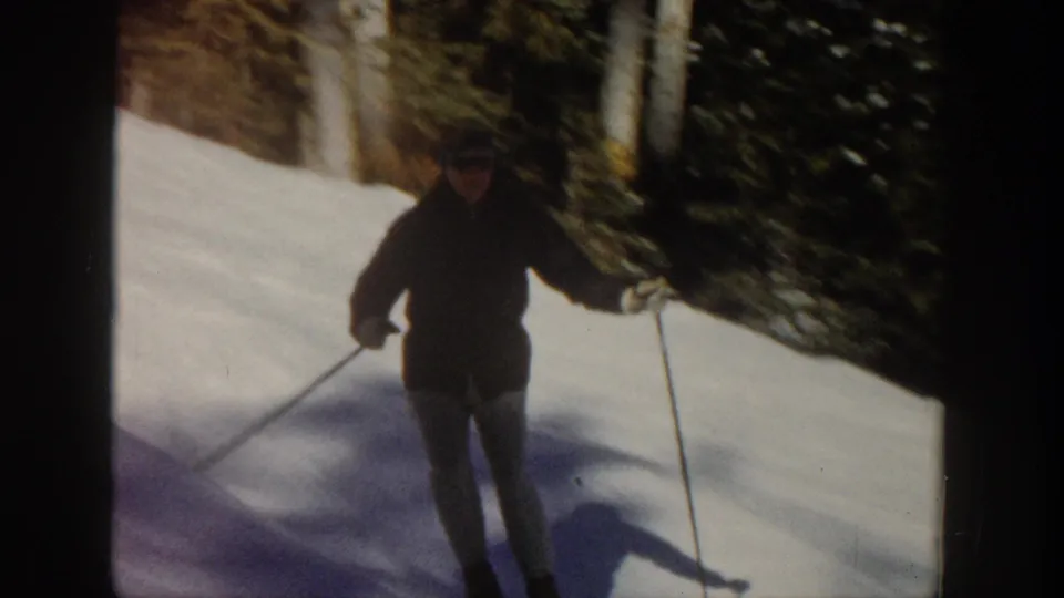 A skier in darker clothing moves across a sunlit Aspen snow slope near evergreen trees in 1961 home-movie footage.