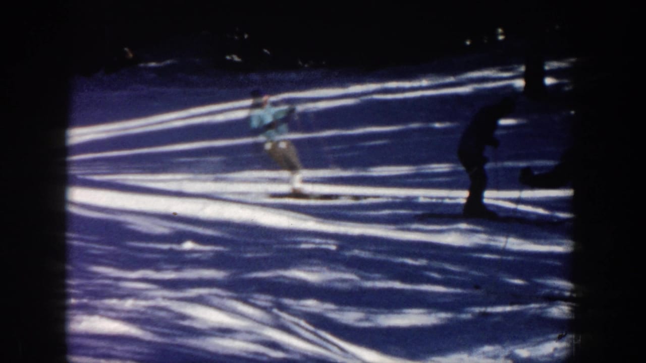 In 1961 Aspen home-movie footage, skiers move downhill across a shadowed snowy slope near evergreen trees.