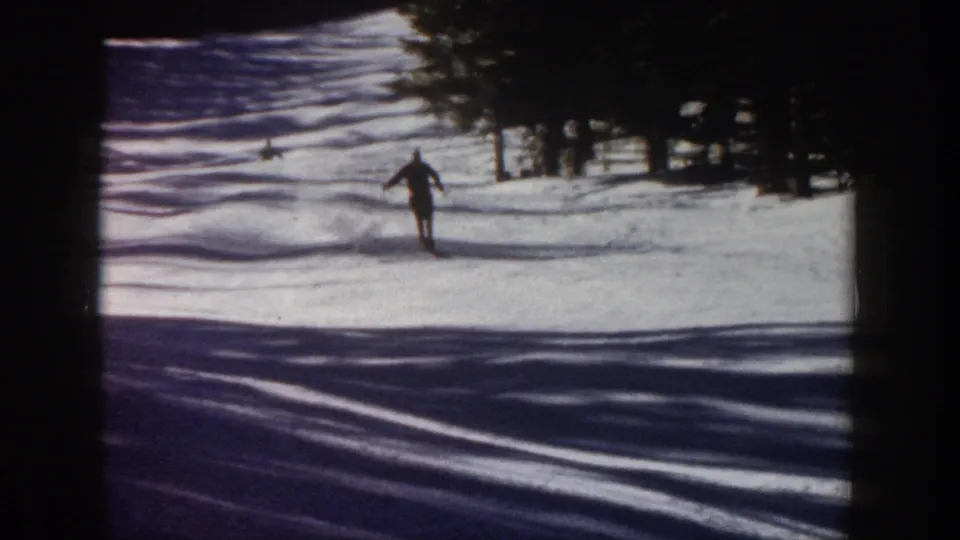 A skier in dark clothing moves toward the camera on a snowy Aspen slope, with trees and shadowed snow behind.