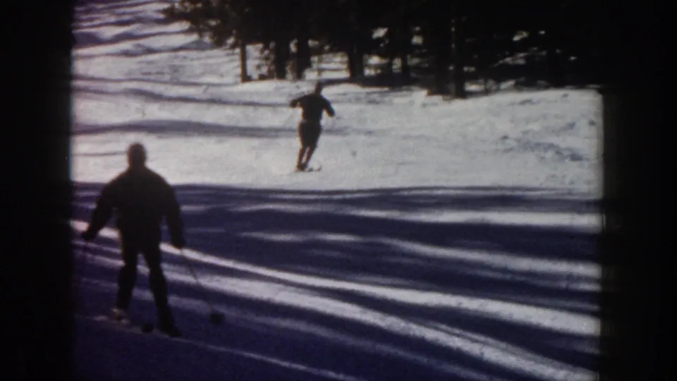 One skier appears near the camera in shadow while another skier descends the snowy Aspen slope beyond.