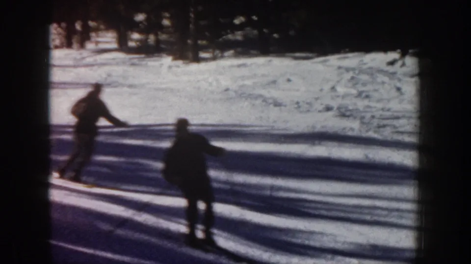 1961 Aspen 8mm frame showing two skiers on a snow-covered downhill area, with pine trees and long shadows behind them.