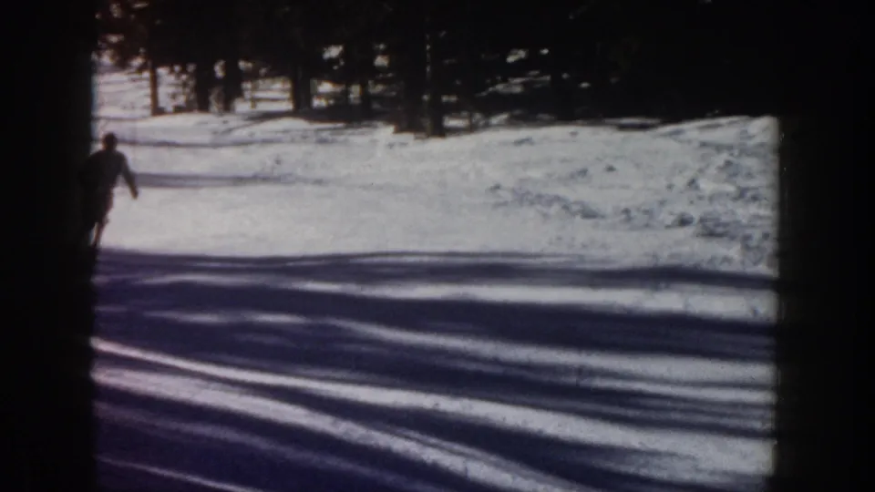 1961 Aspen home-movie frame showing a dark-clothed skier at the left edge of a snowy slope with tree shadows across the snow.