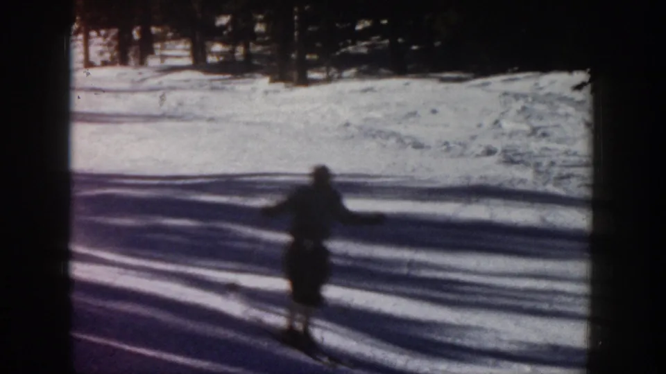 1961 Aspen ski footage frame with a lone skier in dark silhouette crossing bright snow near a line of trees.