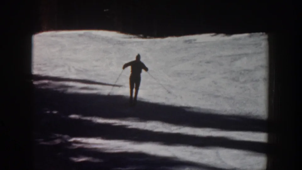 1961 Aspen home-movie frame showing a distant lone skier crossing bright snow under strong backlight and long shadows.