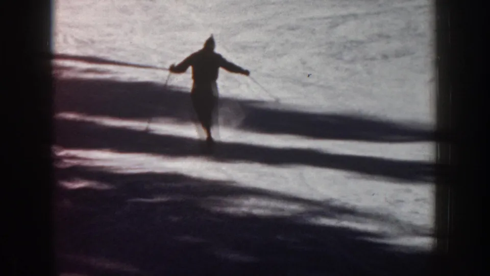 1961 Aspen footage frame with a lone skier in silhouette moving across a sunlit snowy slope with pole lines visible.
