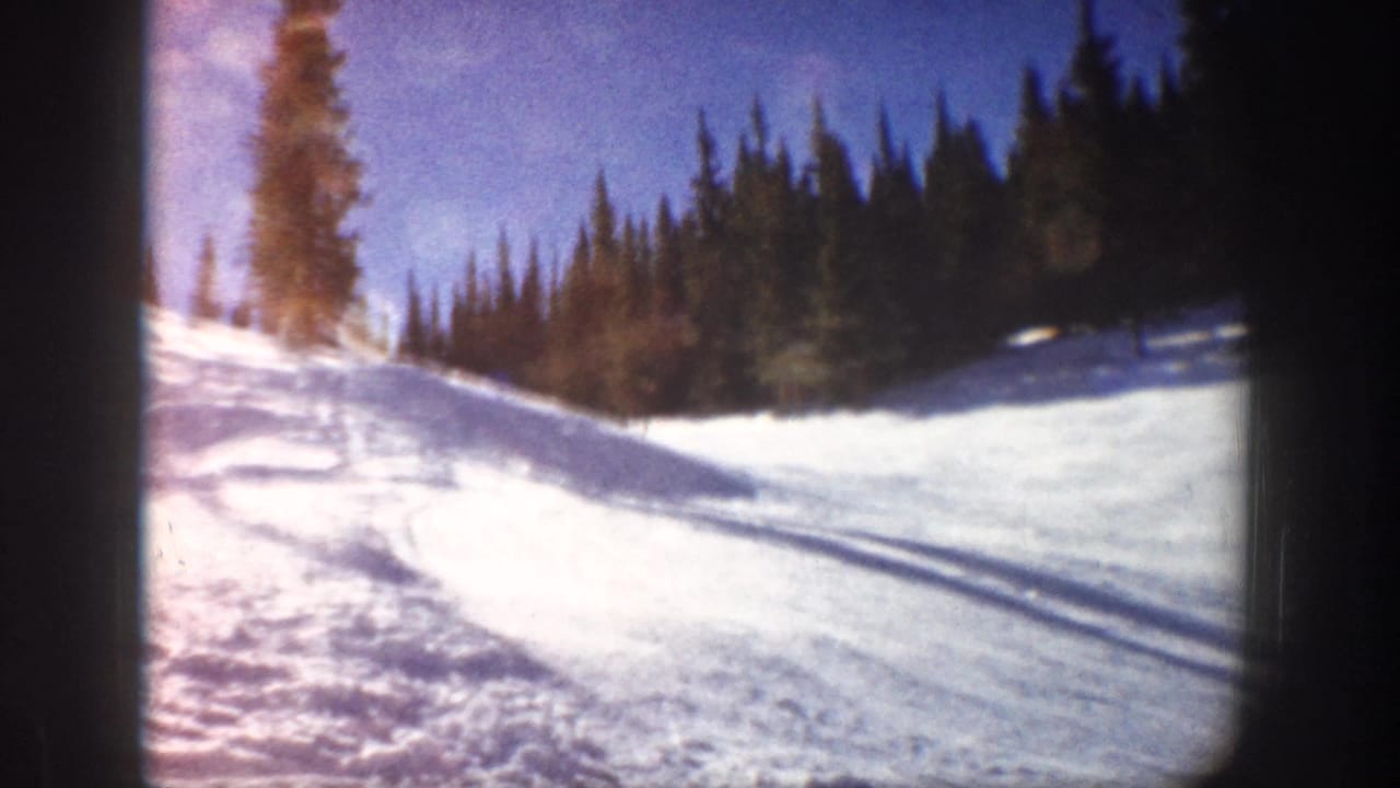 1961 Aspen 8mm frame showing a snowy ski slope bordered by evergreens, with long shadows across the snow.
