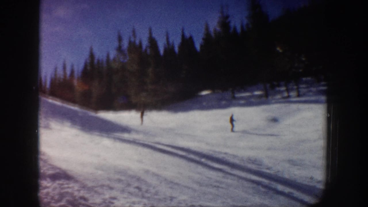 Distant skier crossing a snowy slope below a dark evergreen tree line.