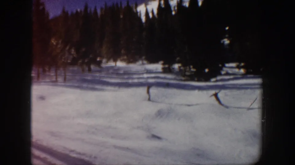 Final 1961 Aspen frame with two distant skiers on bright snow near a dark evergreen stand.