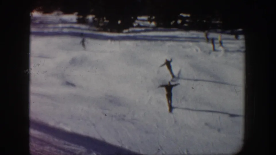 1961 Aspen home-movie frame with a dark-clad skier descending a snowy slope and faint figures near the upper tree line.