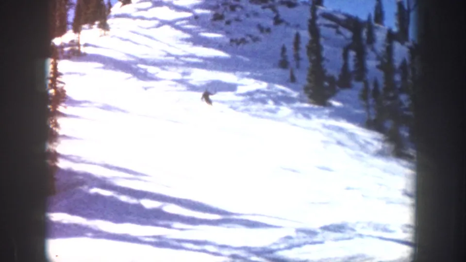 1961 Aspen 8mm frame showing a distant skier on a sunlit snow slope with evergreen trees and strong blue shadows.