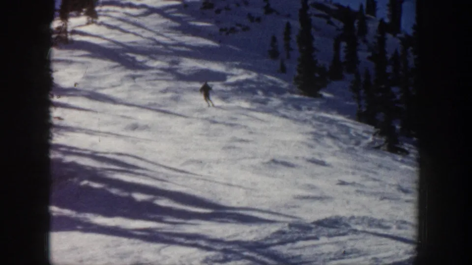 Distant skier on an Aspen, Colorado snowy slope in 1961 home-movie footage, framed by evergreens and long shadows.