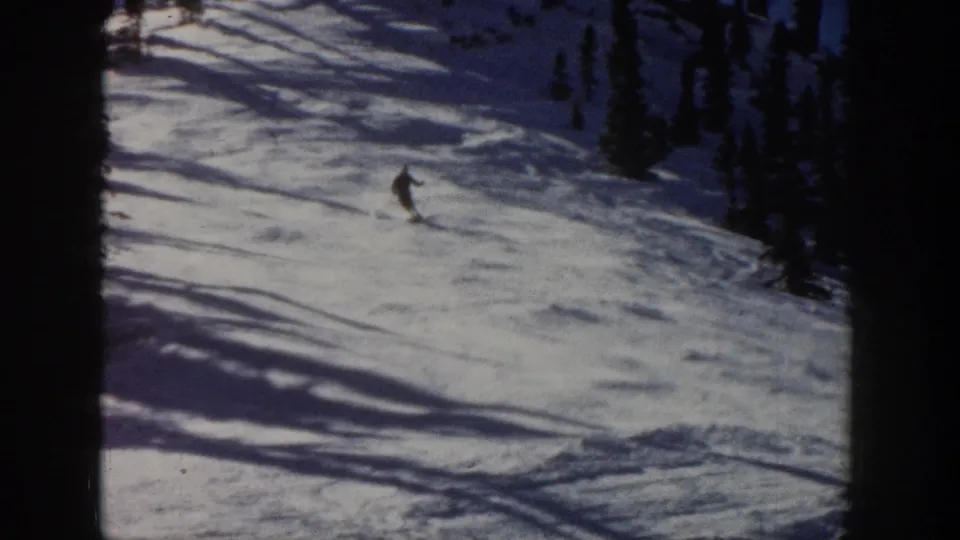 A lone skier turns across the Aspen snowfield, with dark trees and shadow bands visible in the 8mm frame.
