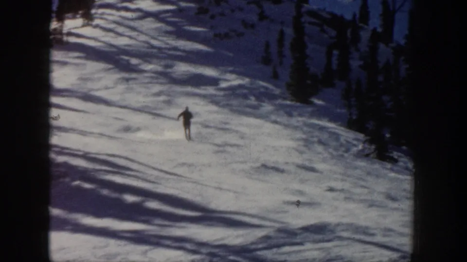 The skier continues downhill on the 1961 Aspen slope, seen small against snow, evergreens, and strong winter shadows.