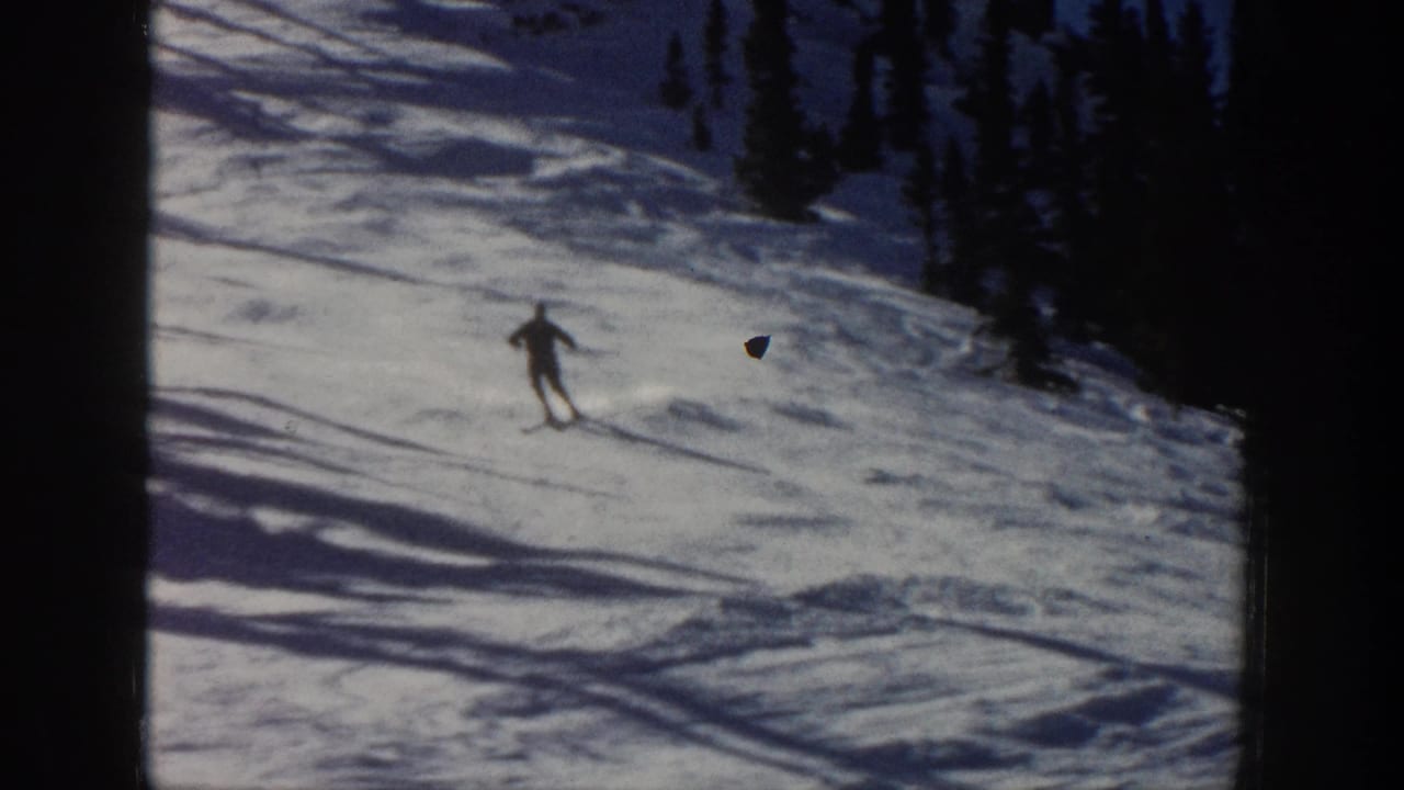 In supported 1961 Aspen footage, a distant skier moves across a snowy mountain slope with trees and blue shadows behind them.