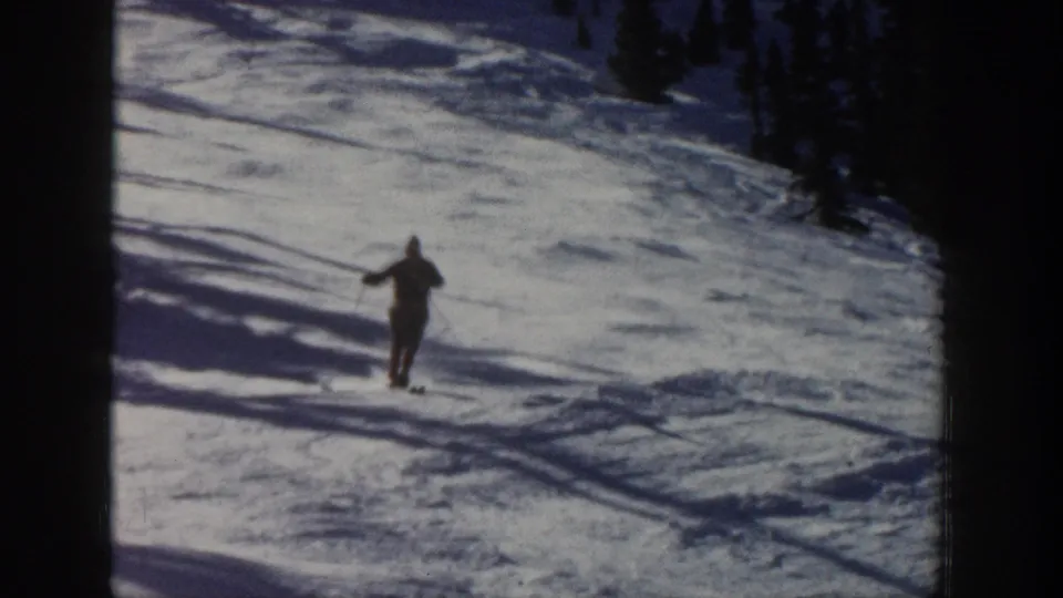 The 1961 Aspen home-movie view shows the skier closer to camera, using poles while crossing tracked snow below evergreens.
