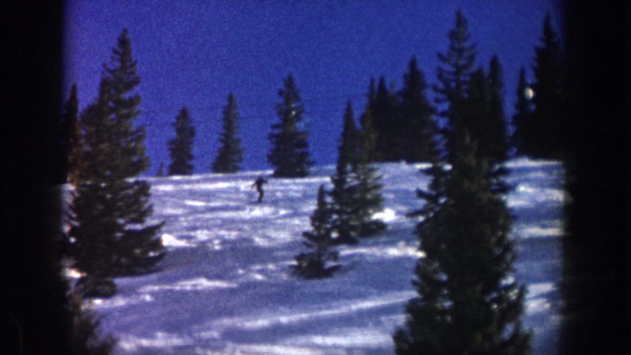 Wide 1961 Aspen winter view with a distant skier crossing a snow-covered slope framed by evergreen trees and blue sky.