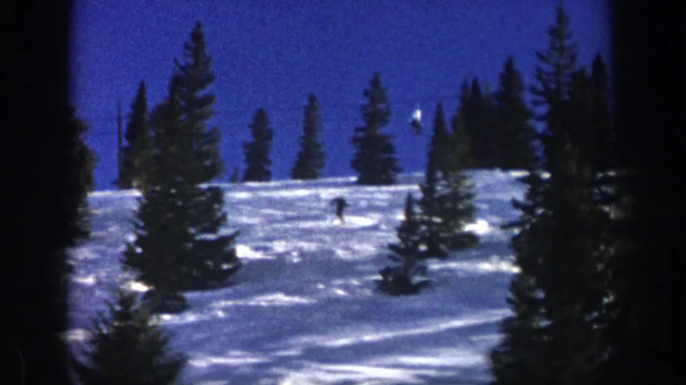 A 1961 Aspen home-movie frame shows the skier small in the landscape, moving across bright snow below tall evergreens.