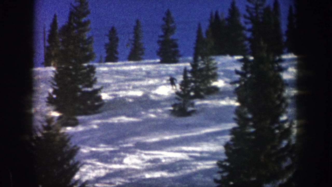 1961 Aspen home-movie frame showing a small skier moving across powder snow among evergreen trees on a bright mountain slope.