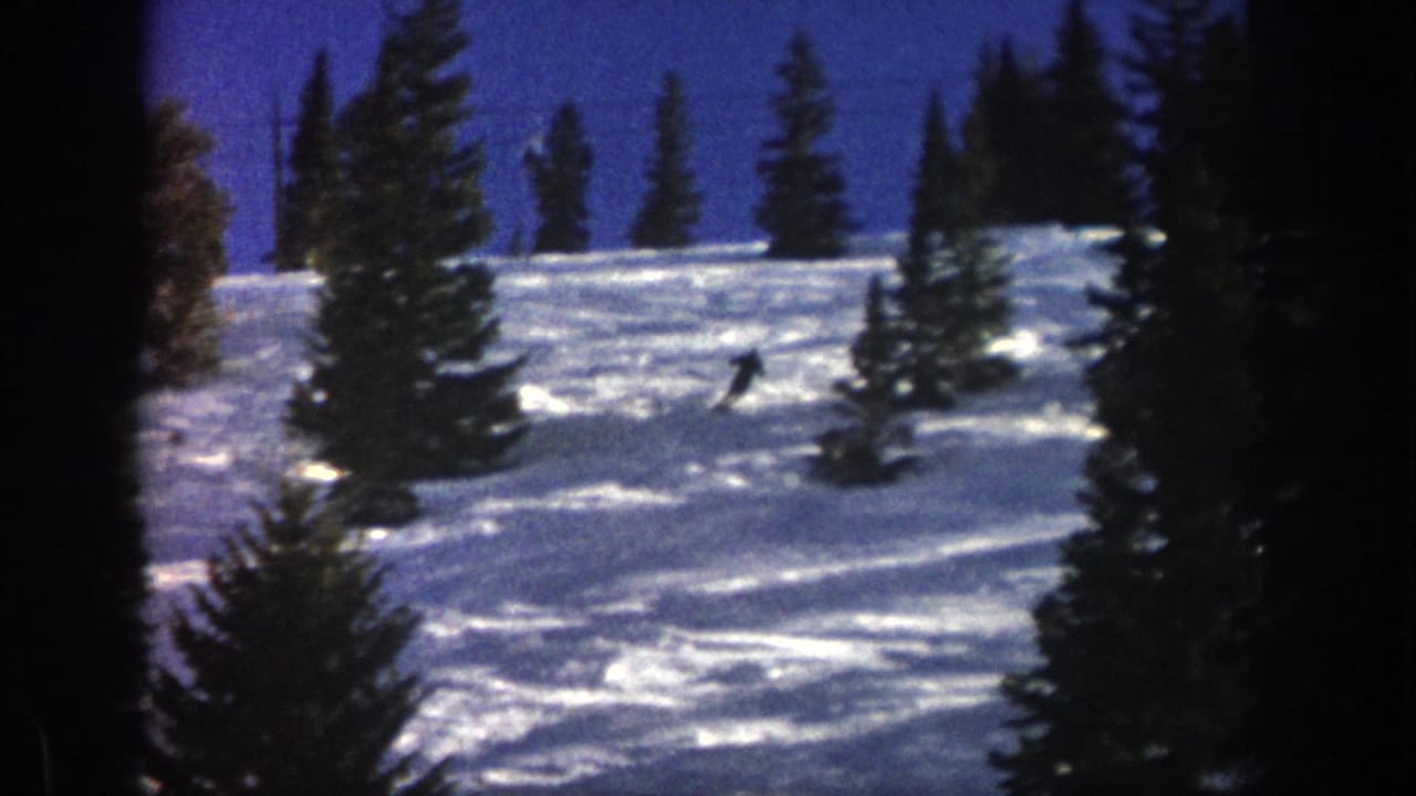 A distant skier crosses an Aspen, Colorado, snowy slope in 1961, framed by dark evergreen trees and saturated blue sky.