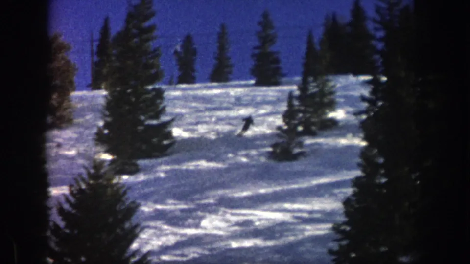 A distant skier crosses an Aspen, Colorado, snowy slope in 1961, framed by dark evergreen trees and saturated blue sky.