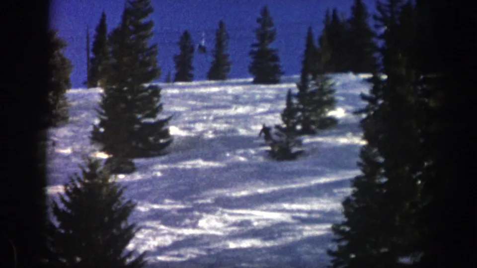 The home-movie frame shows the skier farther downhill on the Aspen slope, with deep snow, evergreens, and dark 8mm frame edges.