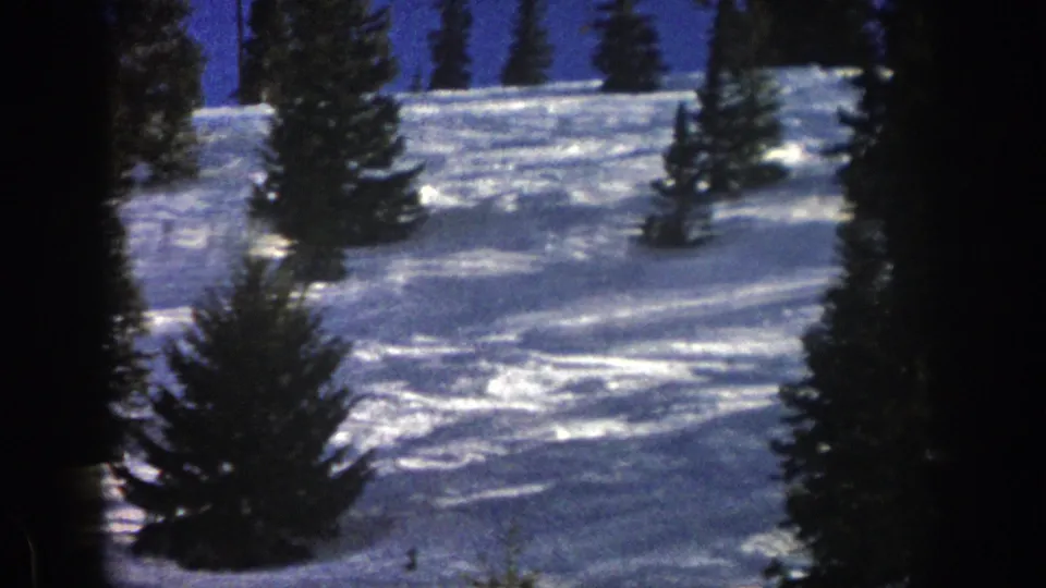 The home-movie view holds on the Aspen ski slope, with evergreens, snow texture, and a distant skier position difficult to distinguish.