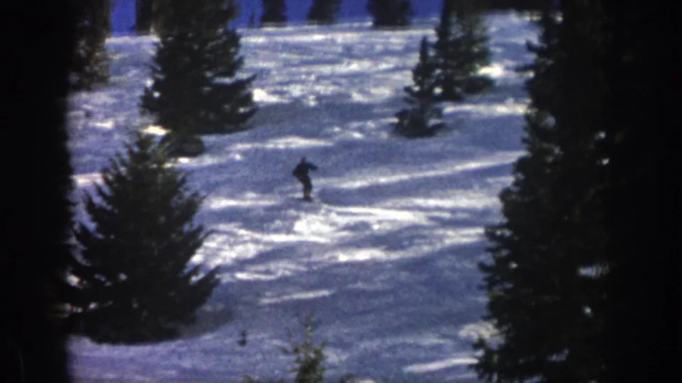 A lone skier in dark winter clothing turns downhill through the snow in the 1961 Aspen home-movie footage.