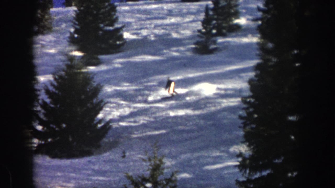 In 1961 Aspen footage, a distant skier in dark winter clothing carves across sunlit snow between tall evergreens.