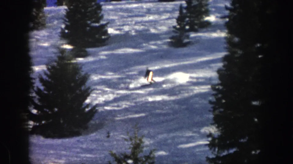 In 1961 Aspen footage, a distant skier in dark winter clothing carves across sunlit snow between tall evergreens.