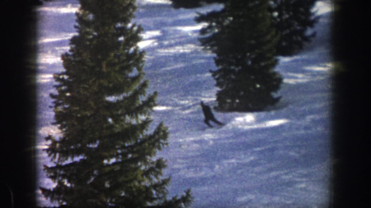 A distant skier in dark clothing crosses an Aspen, Colorado snow slope between tall evergreens in 1961 home-movie footage.
