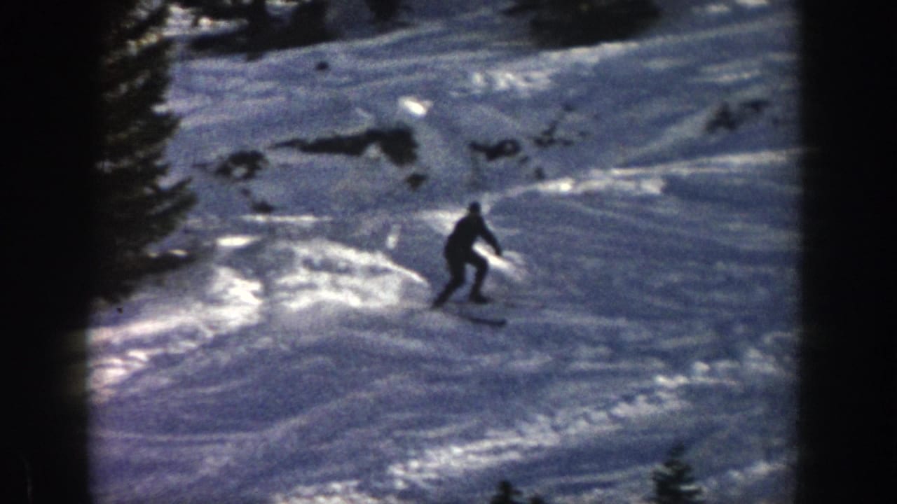 1961 Aspen home-movie frame showing a distant skier moving downhill across bright, uneven snow near dark evergreen trees.