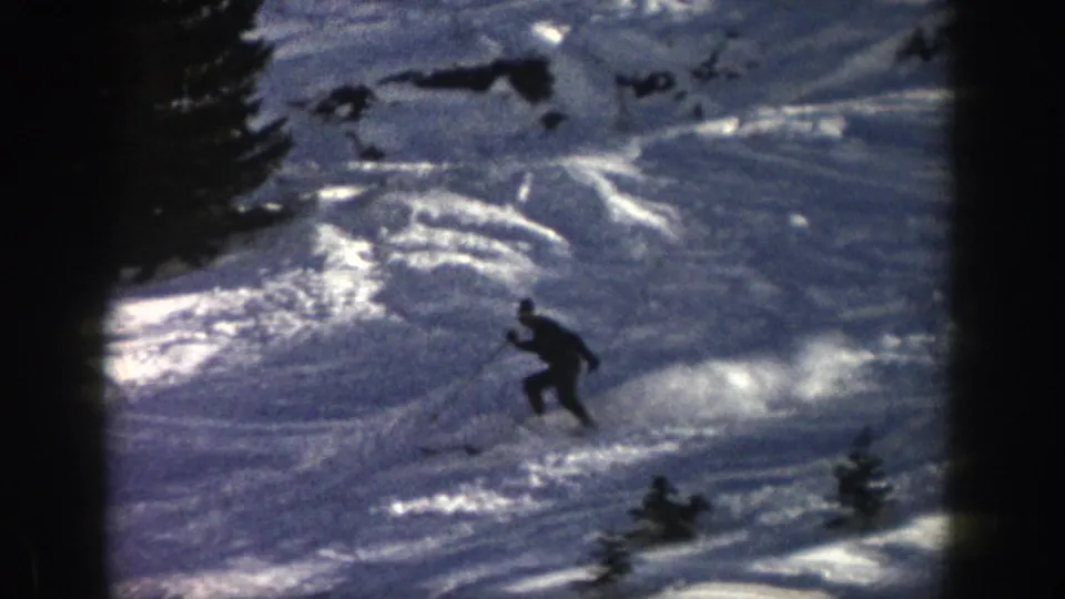 1961 Aspen frame with a distant skier carving across an uneven snowy hillside bordered by dark trees and film-frame shadows.