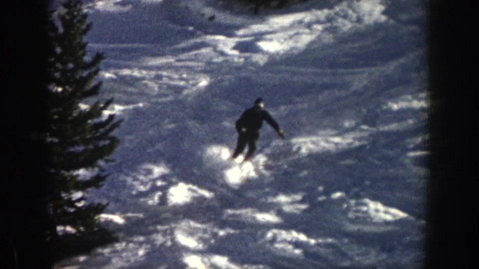 1961 Aspen home-movie still showing the skier lower on the slope, passing through reflective snow with evergreens at the side.