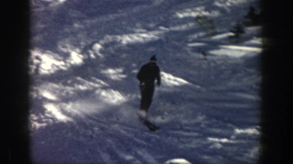 1961 Aspen frame showing the skier from behind on a snowy hillside, with small evergreens and strong reflected winter light.