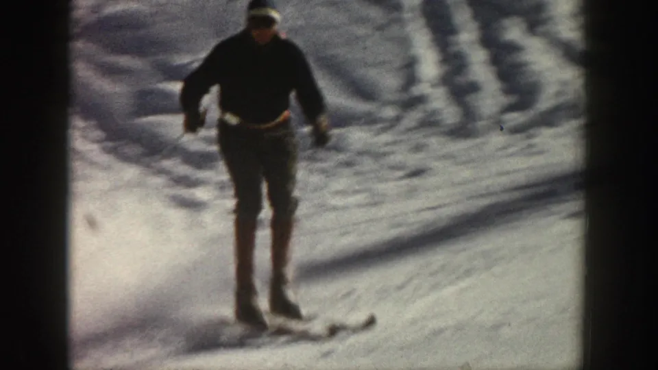 The adult skier approaches in dark winter clothing, using poles as the handheld 8mm camera frames the snowy Aspen slope.