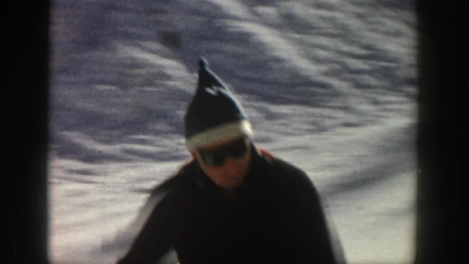 A close handheld frame shows the skier wearing a dark coat, knit cap, and sunglasses against the snowy Aspen background.